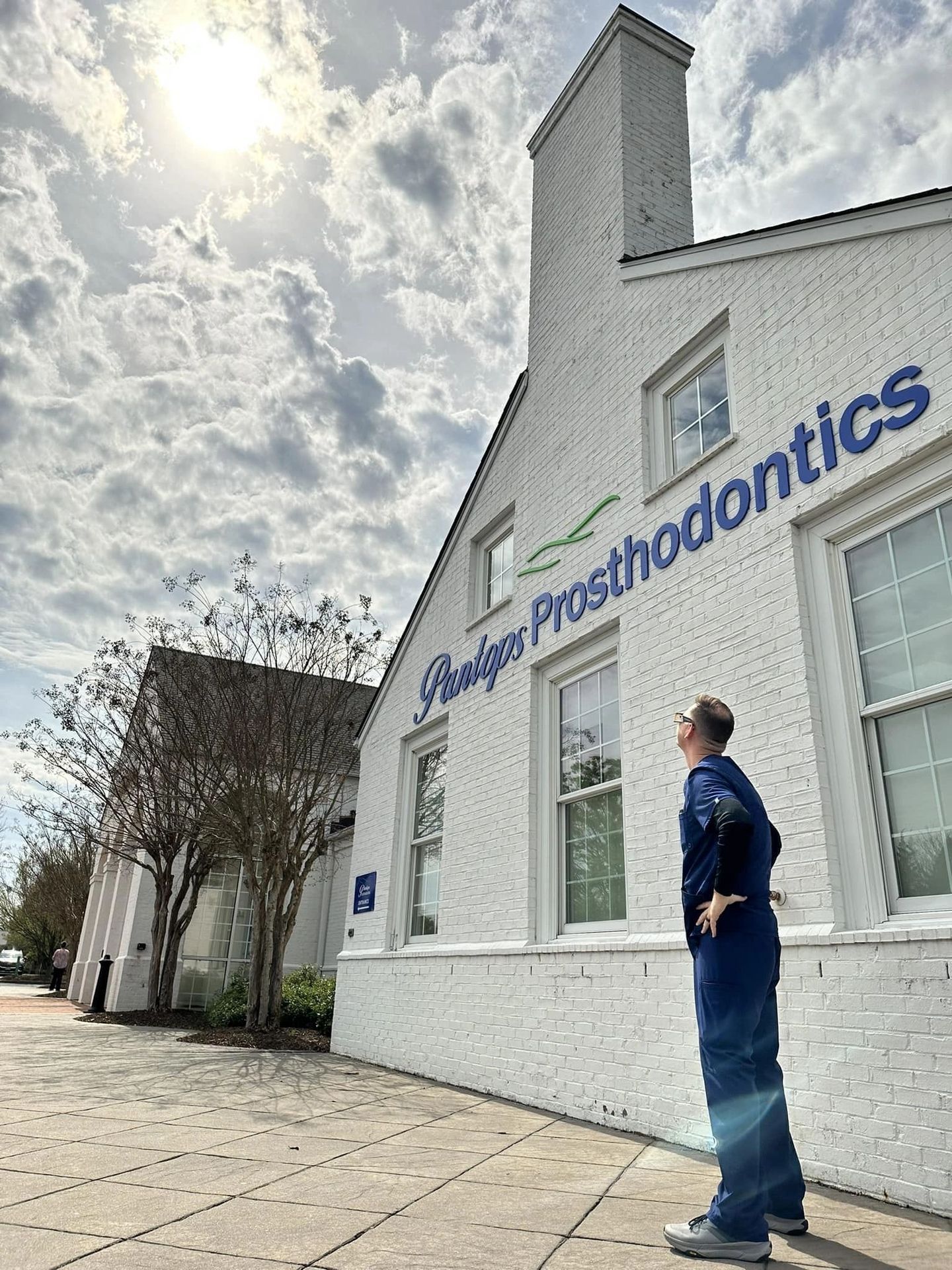 A person in blue scrubs looking up at a Prosthodontics building on a sunny day.